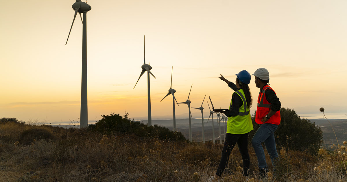 Silhouette of young engineer holding laptop computer planning and working for the energy industry and standing beside a wind turbines farm power station at sunset time