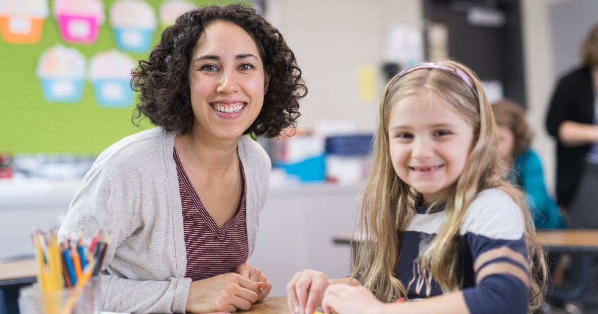 A first grader works on her math sheet at her desk. Her teacher is sitting next to her helping her out with a challenging problem on it. They're both looking up at the camera and smiling.