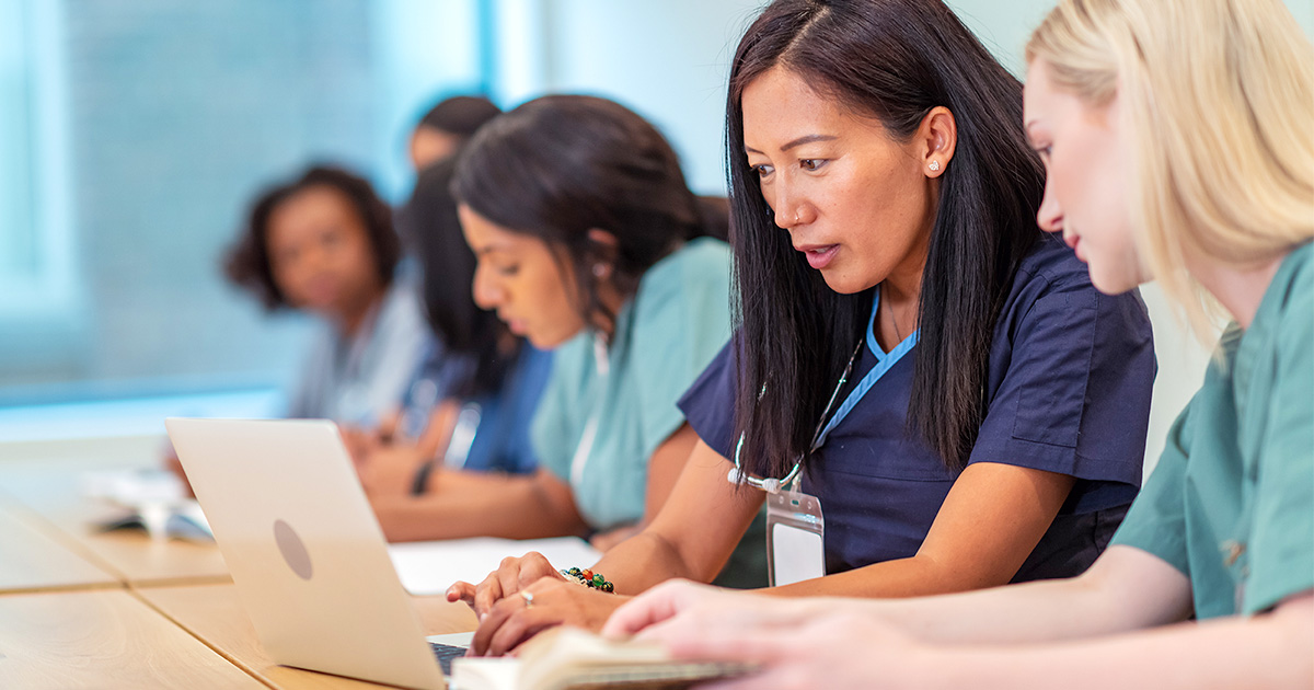 A group of focused medical professionals in scrubs collaborate at a table. A woman in the foreground types on a laptop, conveying teamwork and study.