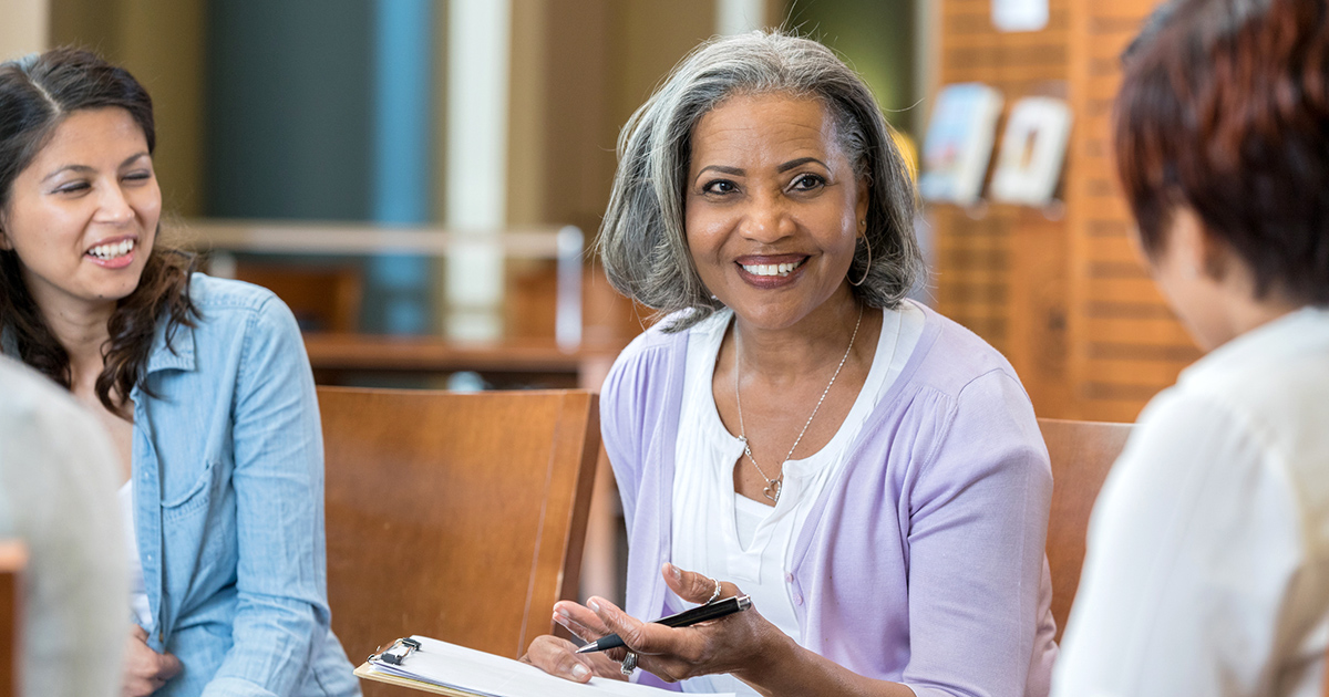 A senior female university professor sits in a circle with her class in the library.  She holds a clipboard as she speaks to the group.