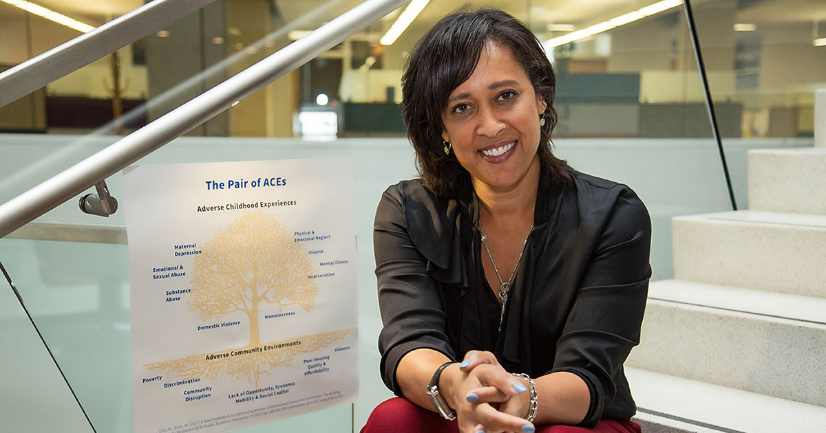 Dr. Wendy Ellis, smiling at the camera while sitting on a staircase next to a poster with an illustration titled "The Pair of ACEs: Adverse Childhood Experiences".