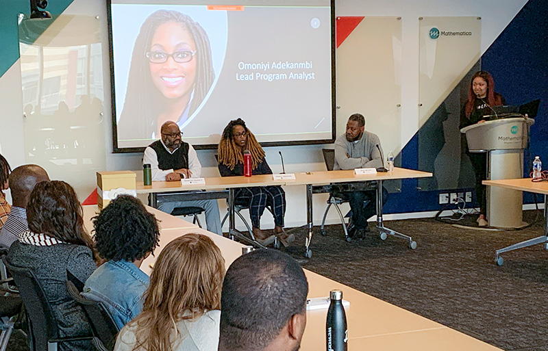 Demetrius Goosbey, Omoniyi Adekanmbi and Derek Mitchell speaking to a group in the D.C. office with a presentation behind them. 