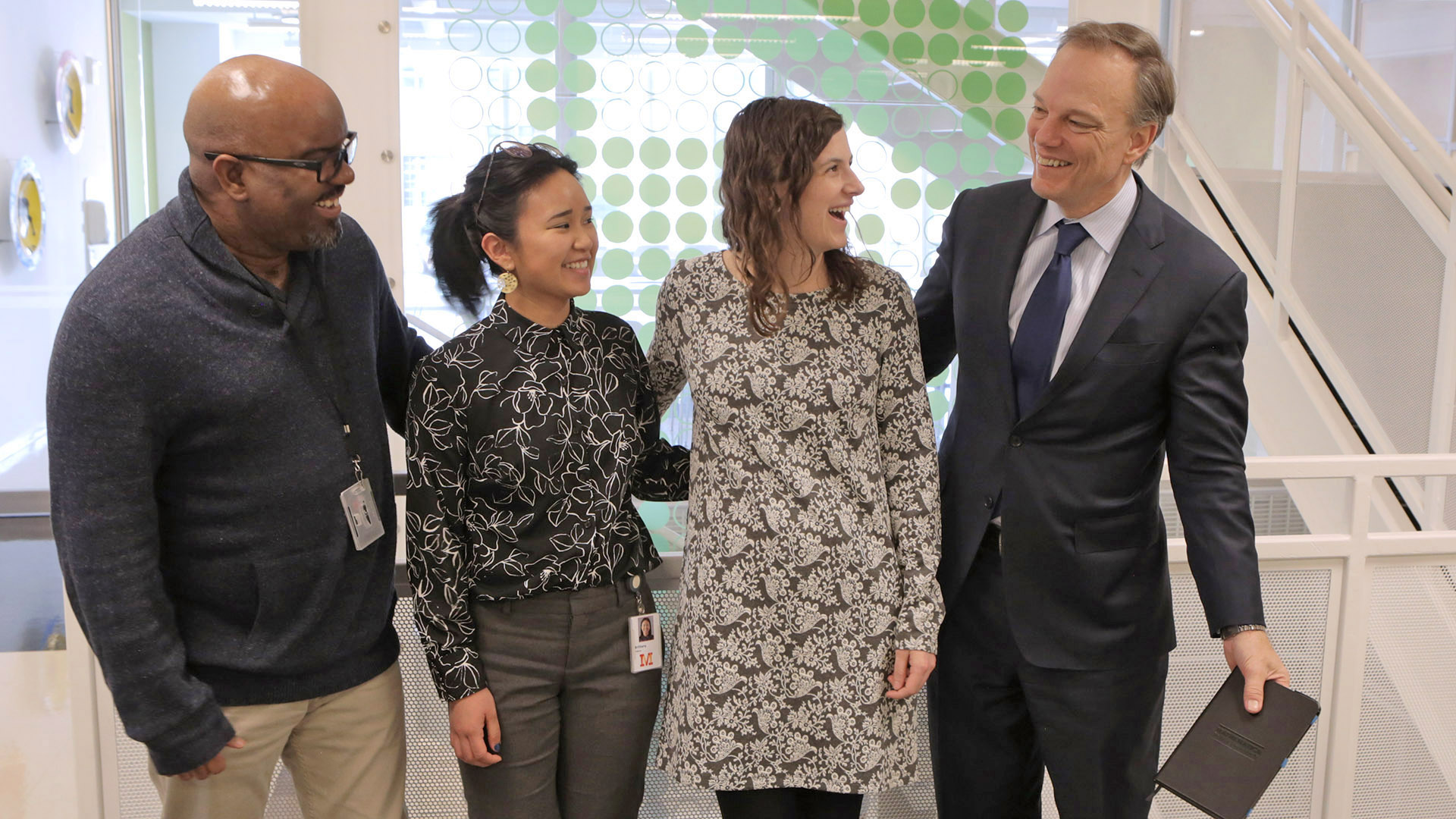 Four people, including Paul Decker and Demetrius Goosbey, standing in a row in the Mathematica DC office, with arms around each other, smiling at each other. 