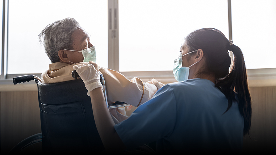 Nurse taking care of mature male patient sitting on wheelchair in hospital, both wearing masks