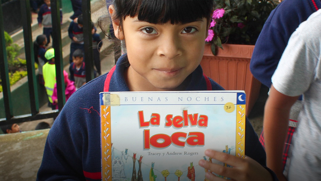 Child facing camera and smiling shyly, holding up book with title: La selva loca
