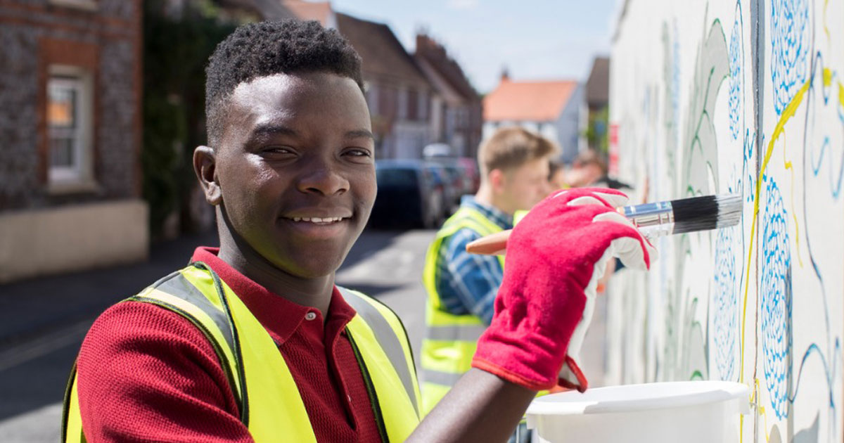 Teen in a work vest painting a mural on a wall. 