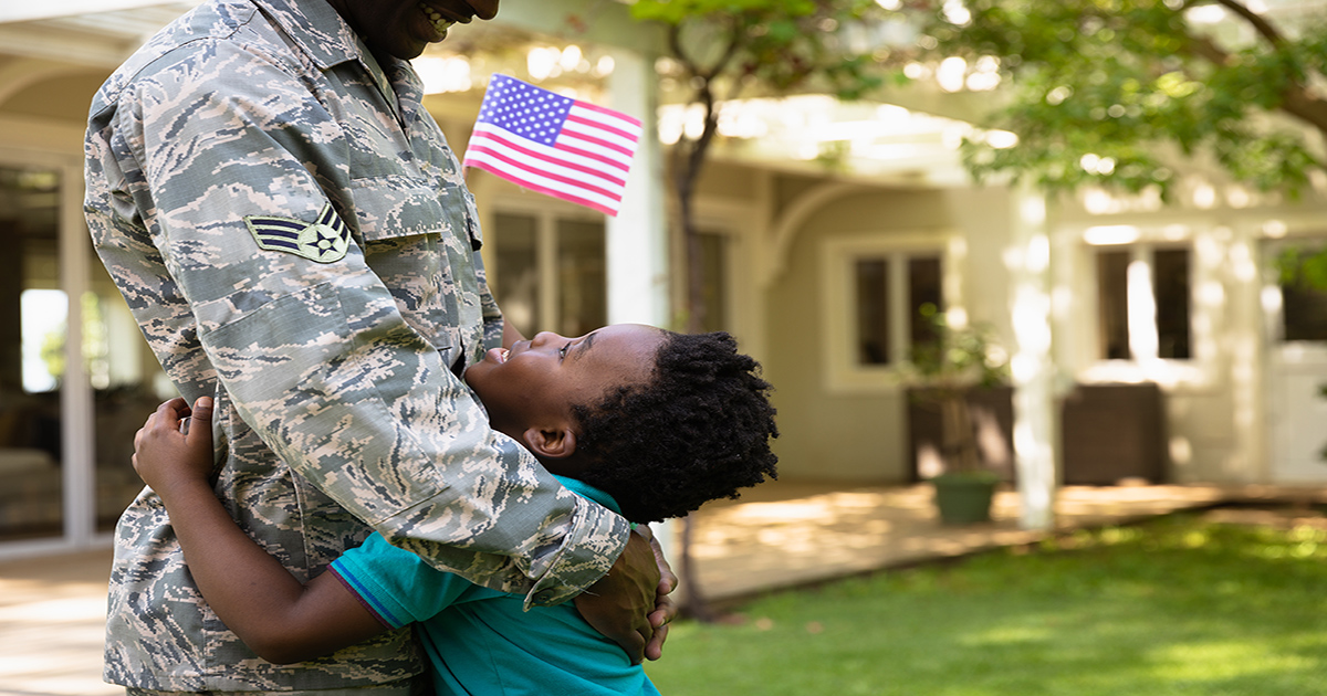 Side view close up of a young adult African American male soldier in the garden outside his home, embracing his young son, who is looking up at him smiling and holding a US flag