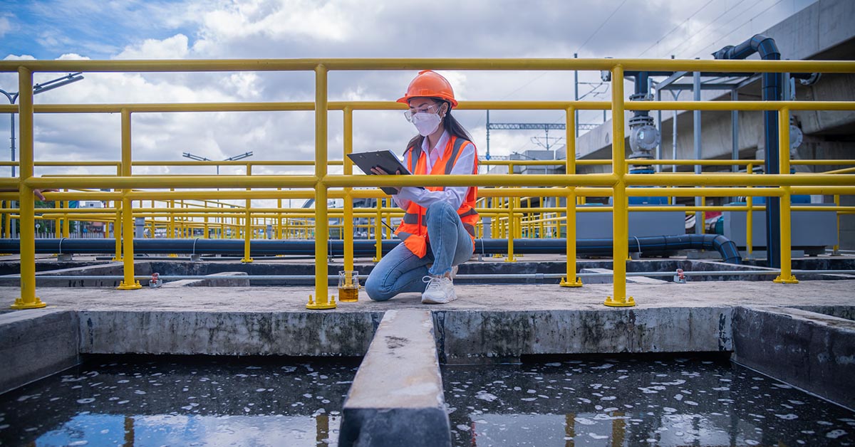 A worker takes water from a wastewater treatment pond to check the quality of the water. She wears a face mask to protect herself from pollution.