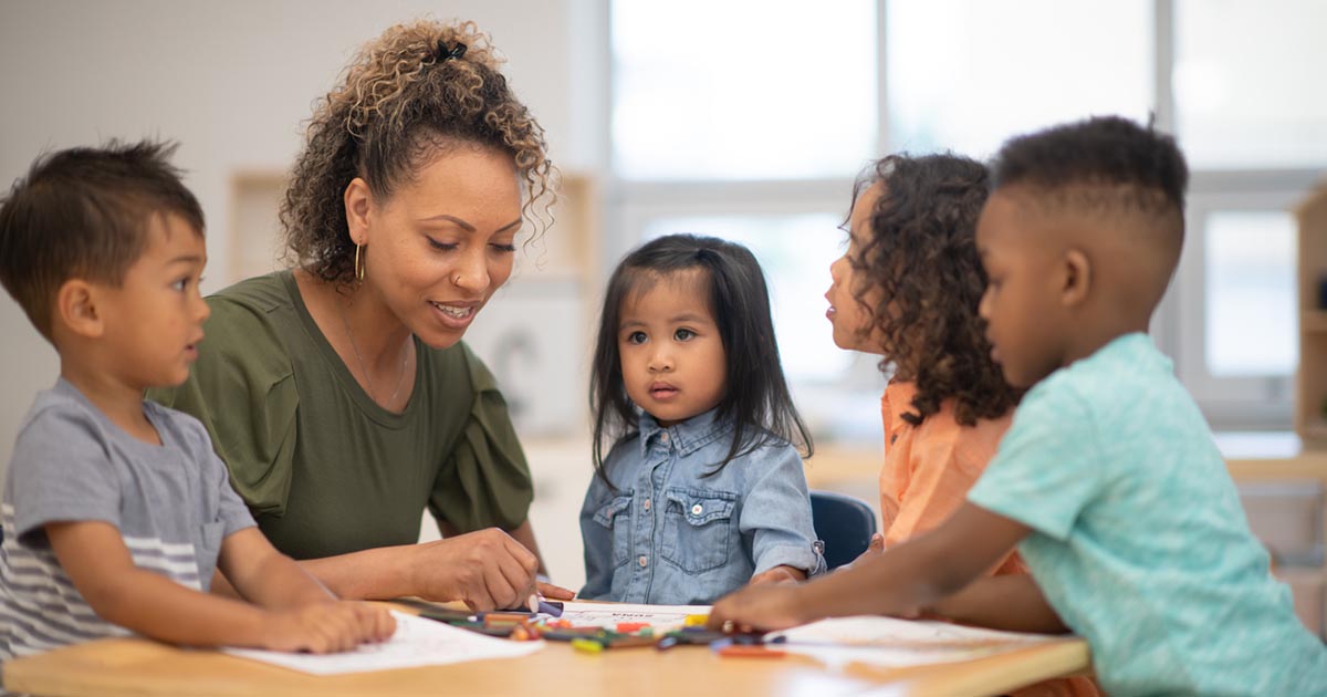 A multi ethnic group of preschool children are colouring together at a desk with their preschool teacher.