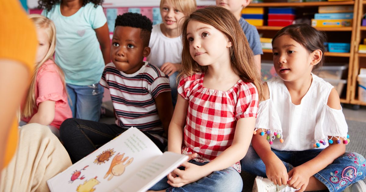 Group Of Elementary School Pupils Sitting On Floor Listening To Female Teacher Read Story