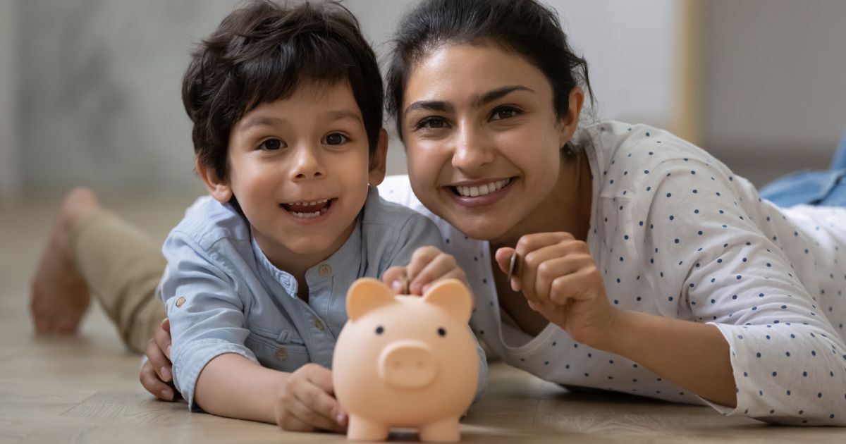 Portrait of happy indian ethnic family lying on floor with small piggybank. Caring young asian mum teaching little child son saving money for future or planning purchases, financial education concept.