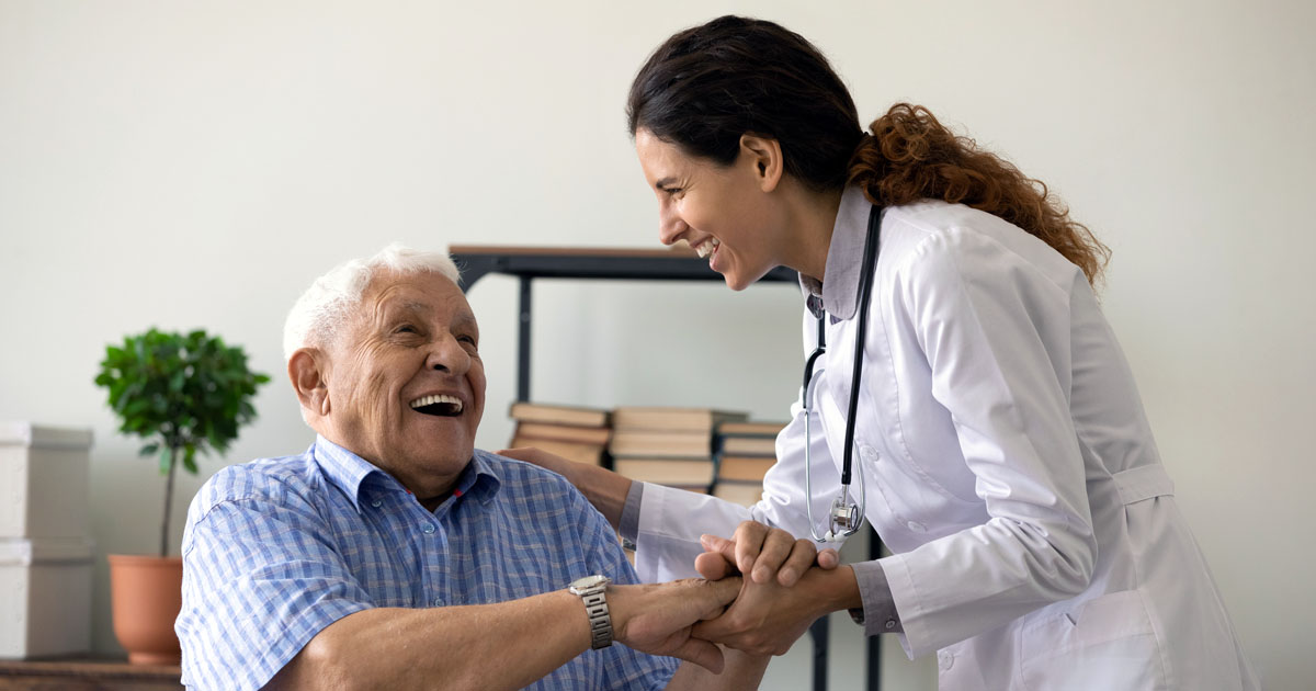 A smiling elderly man grasps the hands of a medical professional in an office. 