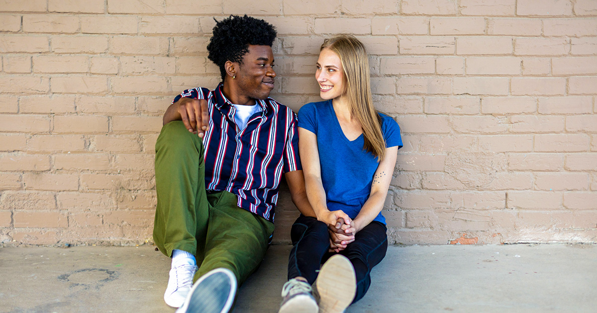 Smiling young couple sitting together against a brick wall, holding hands and looking at each other.