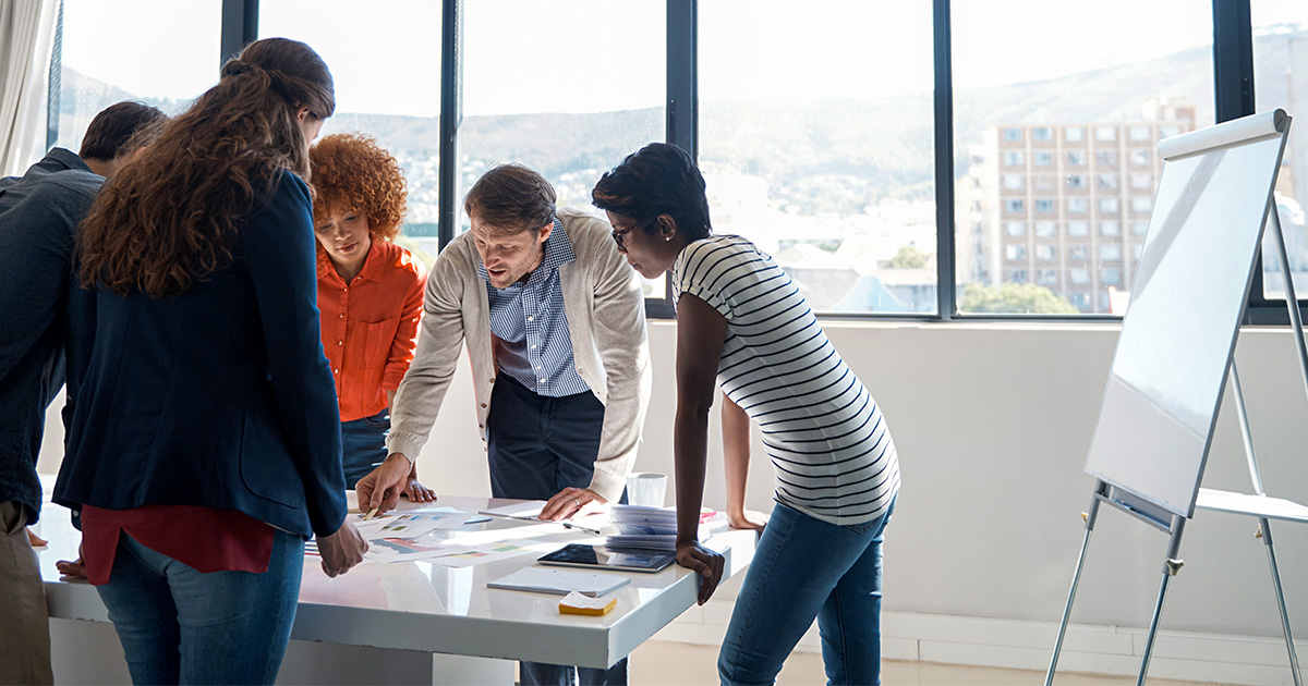 A group of five people leans over a table in a brightly lit room, collaborating on documents. They appear engaged and focused, with an empty easel nearby.