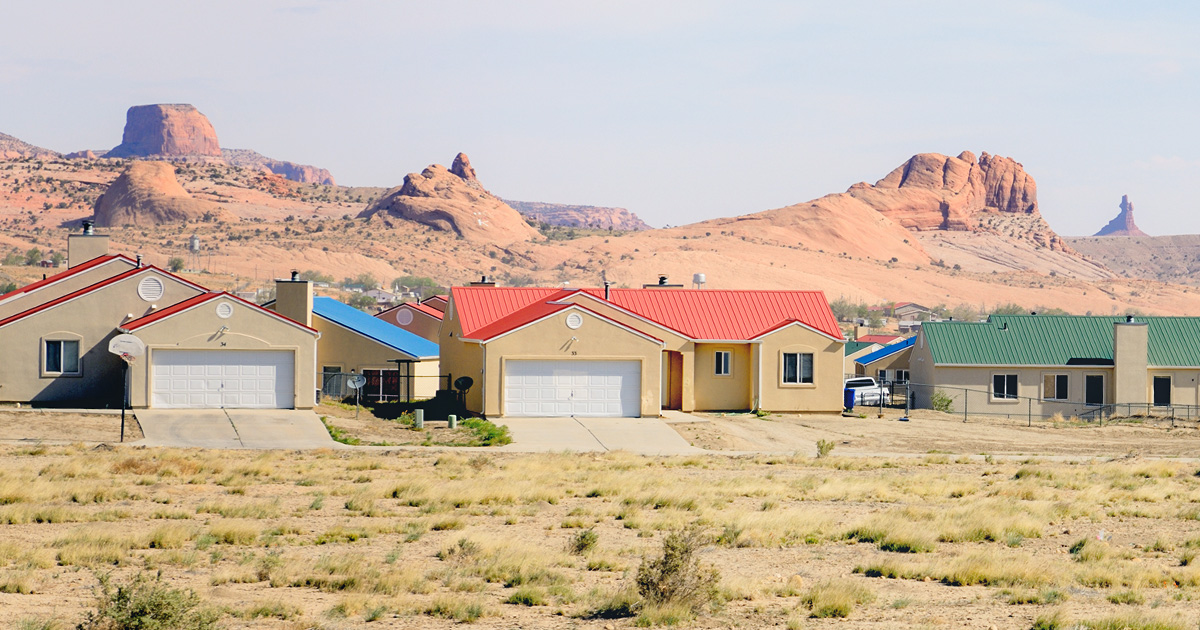 A suburban neighborhood with colorful roofs sits in a desert landscape. Sparse vegetation covers the ground, and distant rocky formations rise under a clear sky.