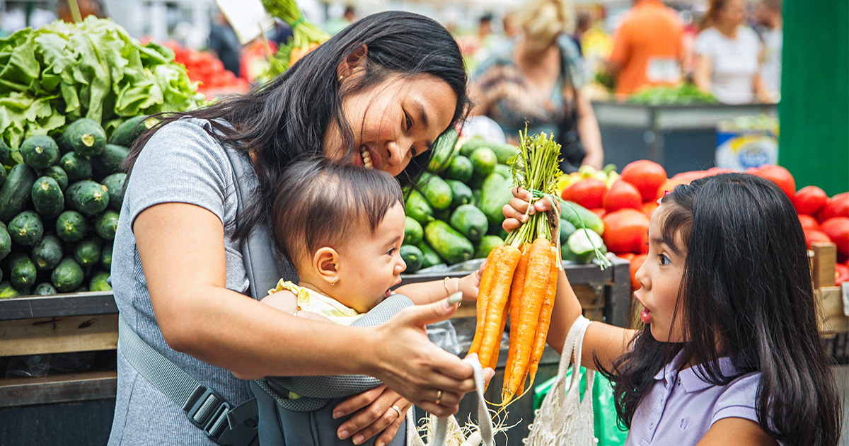 A mother and two children happily shop for vegetables at a market, surrounded by fresh produce