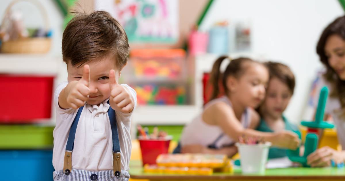 A toddler looks into the camera and gives two thumbs up while classmates play behind them