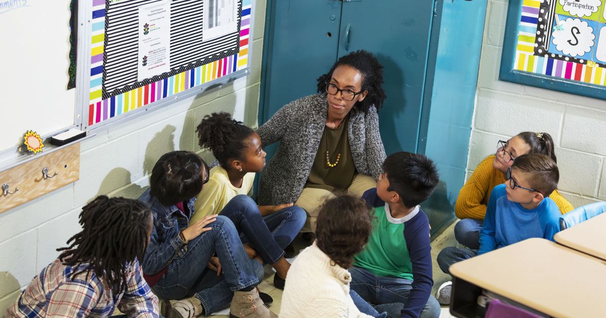 A multiracial group of seven school children and their teacher doing a practice drill, sheltering in place, sitting on the floor in a corner of their classroom.