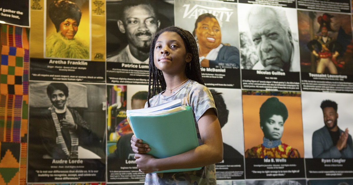 A seventh-grade Black girl walks in front of a display of famous Black Americans, including Audre Lorde, Aretha Franklin, and Ryan Coogler, at Sutton Middle School.  